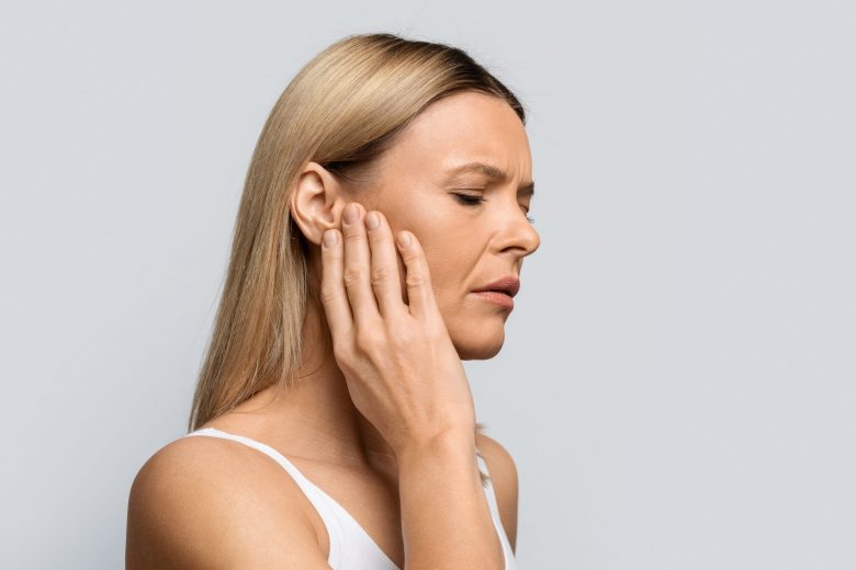 Tinnitus. Profile of sick female middle aged blonde woman having strong ear pain, touching her painful head, isolated on grey studio background, panorama with copy space